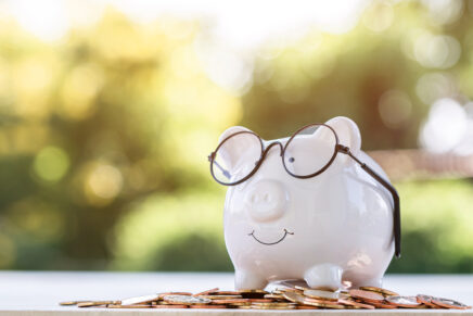 Piggy bank and large sums of money placed on a wooden table in the garden next to the house. Financial planning for future use and emergencies or investing for profit concept.